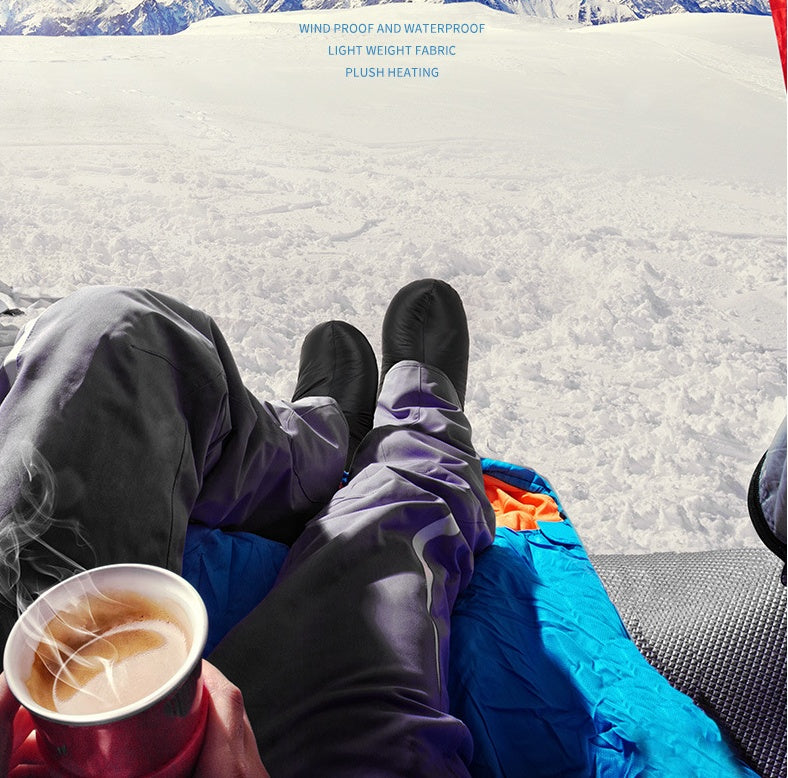 Person sitting on a sleeping bag in the snow holding a cup of coffee.