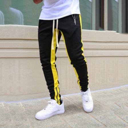 Person wearing black jogger pants with yellow stripes and white sneakers on a concrete surface.