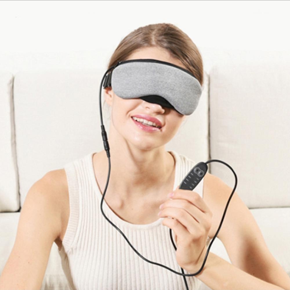 Woman wearing a gray eye mask with a remote control, sitting on a beige couch.