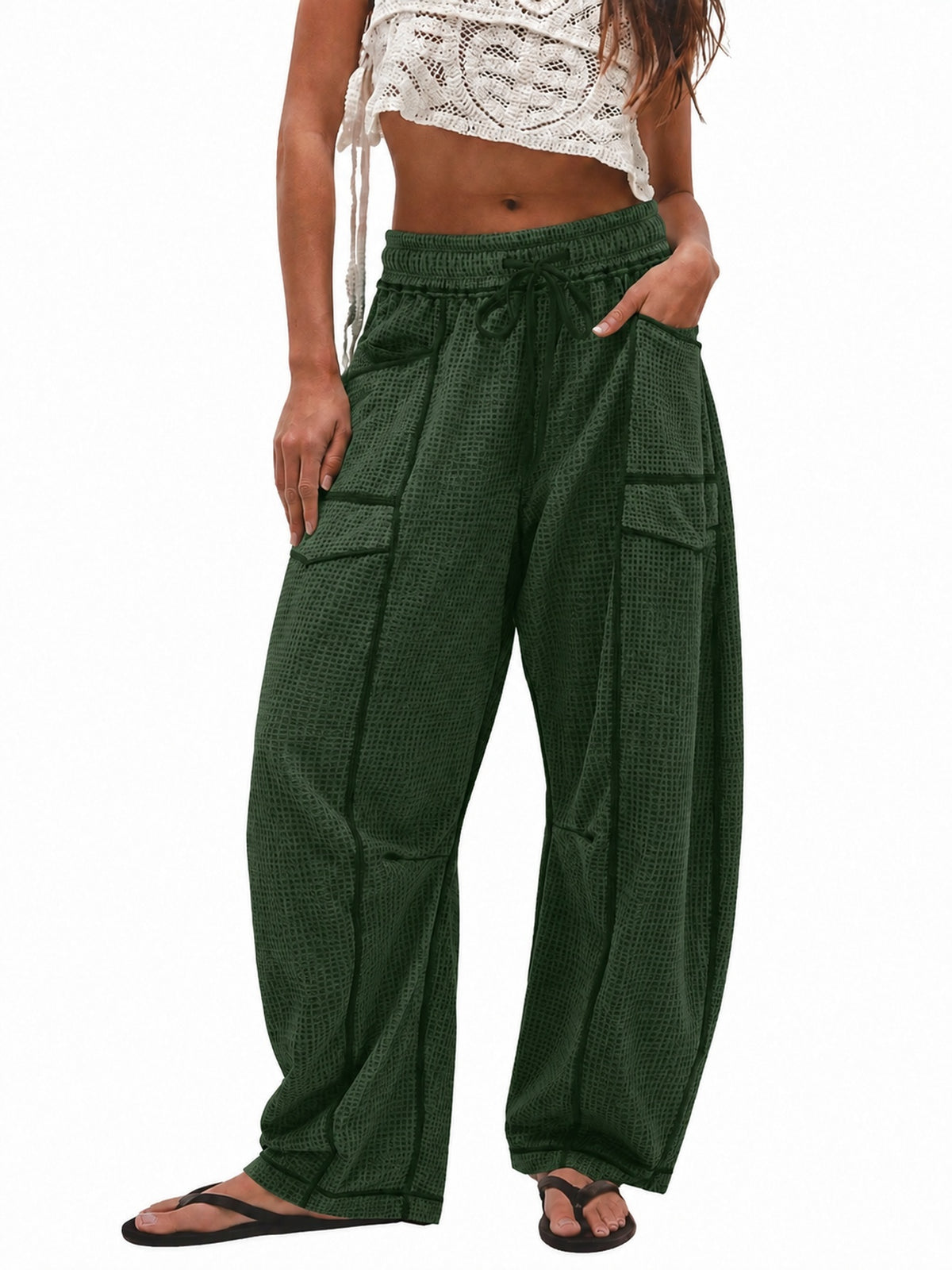 A model wearing dark green wide-leg cargo pants with a textured pattern, paired with a white crochet crop top and black flip-flops, shown against a plain white background.