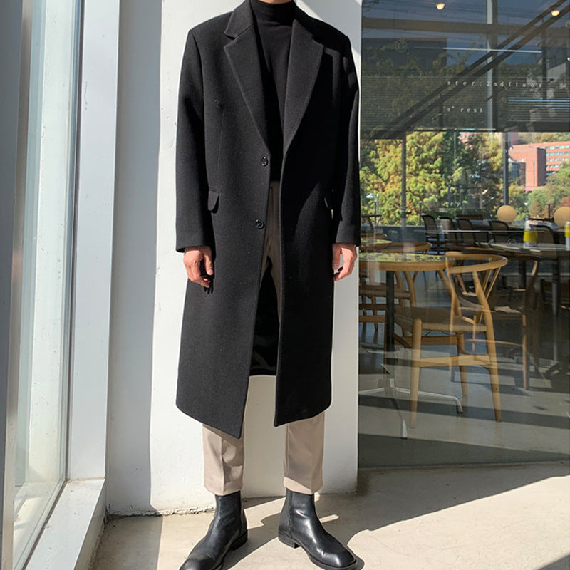 A person wearing a long black overcoat over a black turtleneck, beige trousers, and black ankle boots, standing in front of a glass storefront window.