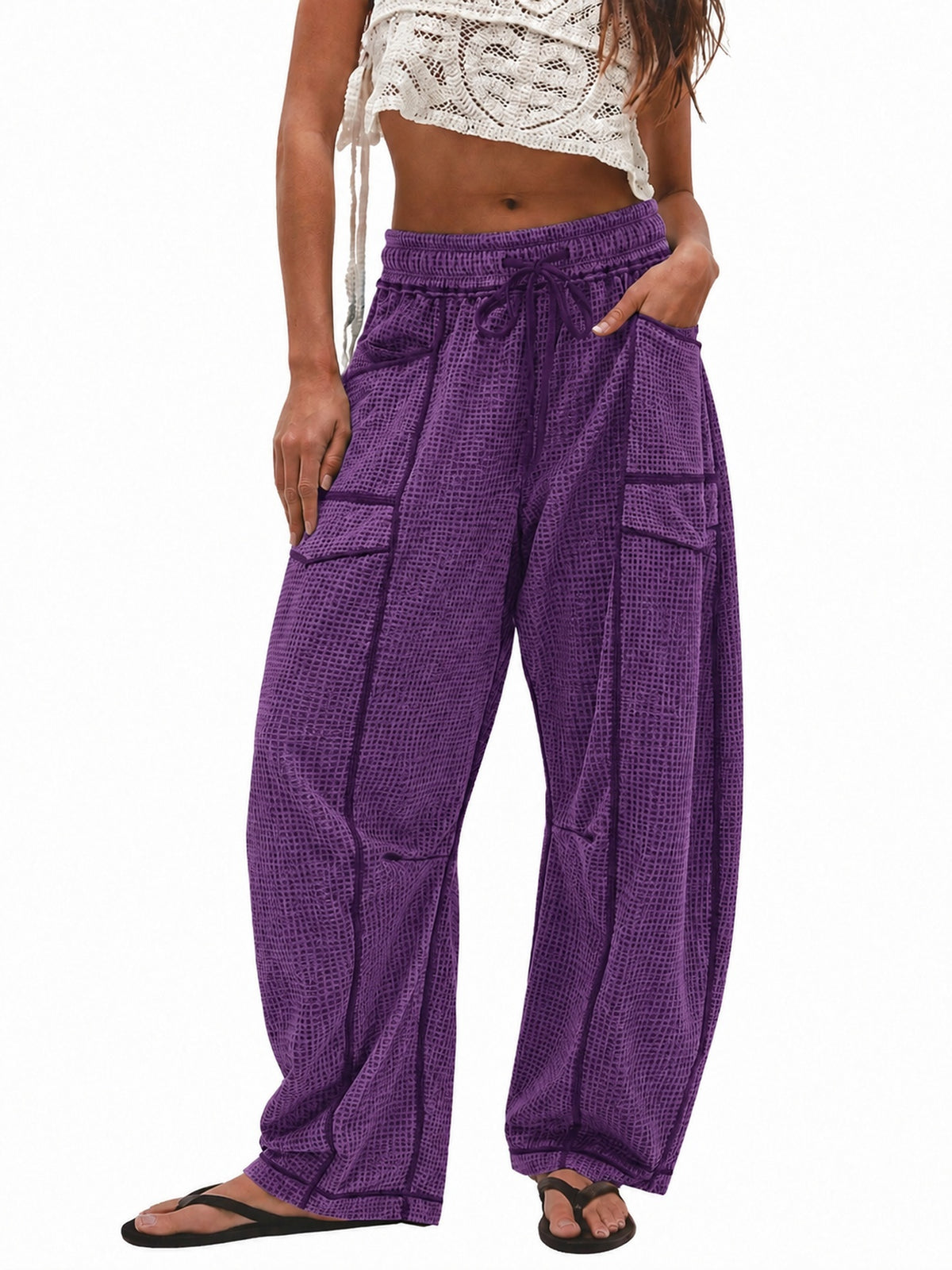 A model wearing purple harem-style pants with a subtle grid pattern, featuring a drawstring waist, side pockets, and a relaxed fit, paired with a white crochet crop top and black flip-flops against a white background.