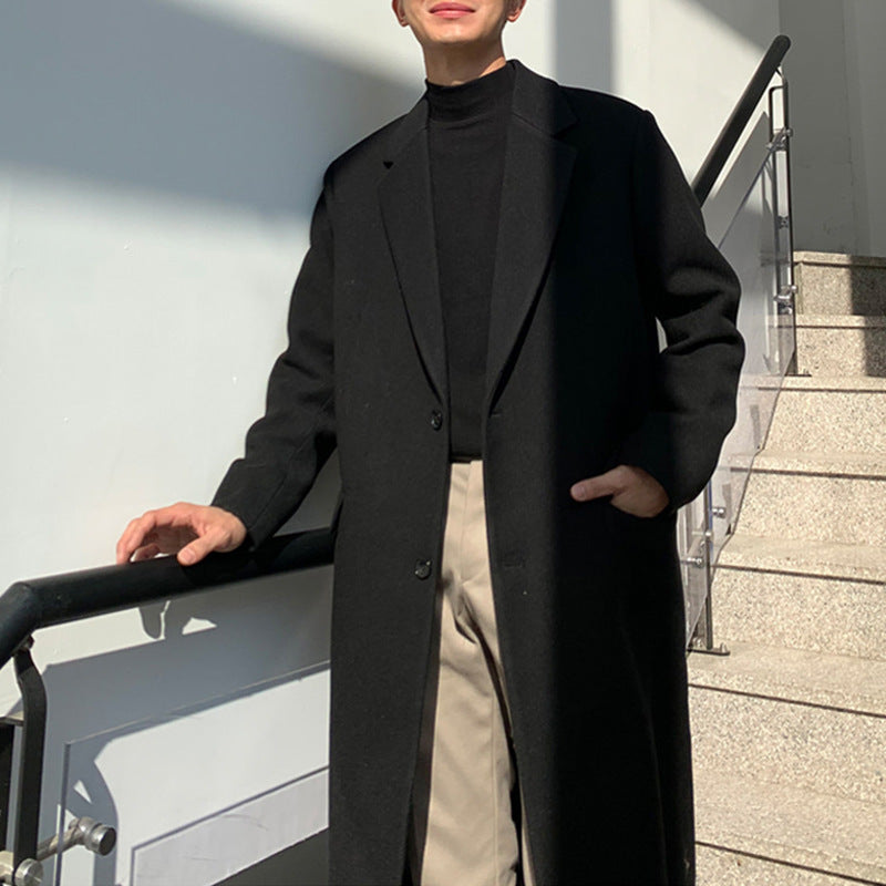 A person wearing a long black overcoat over a black turtleneck and beige trousers, standing beside an outdoor staircase with a metal railing.