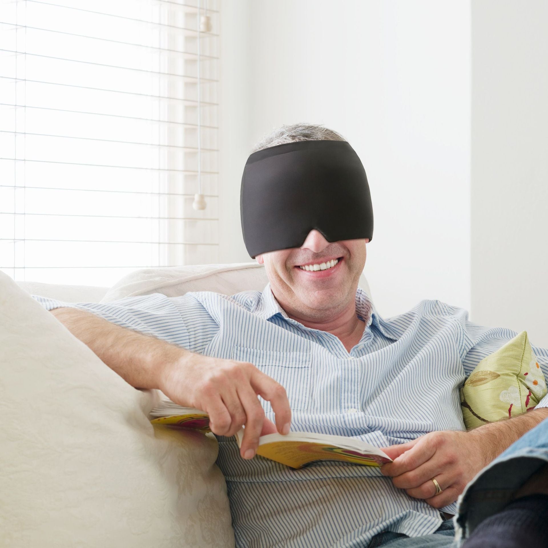 Man wearing a black sleep mask while reading a book on a couch.