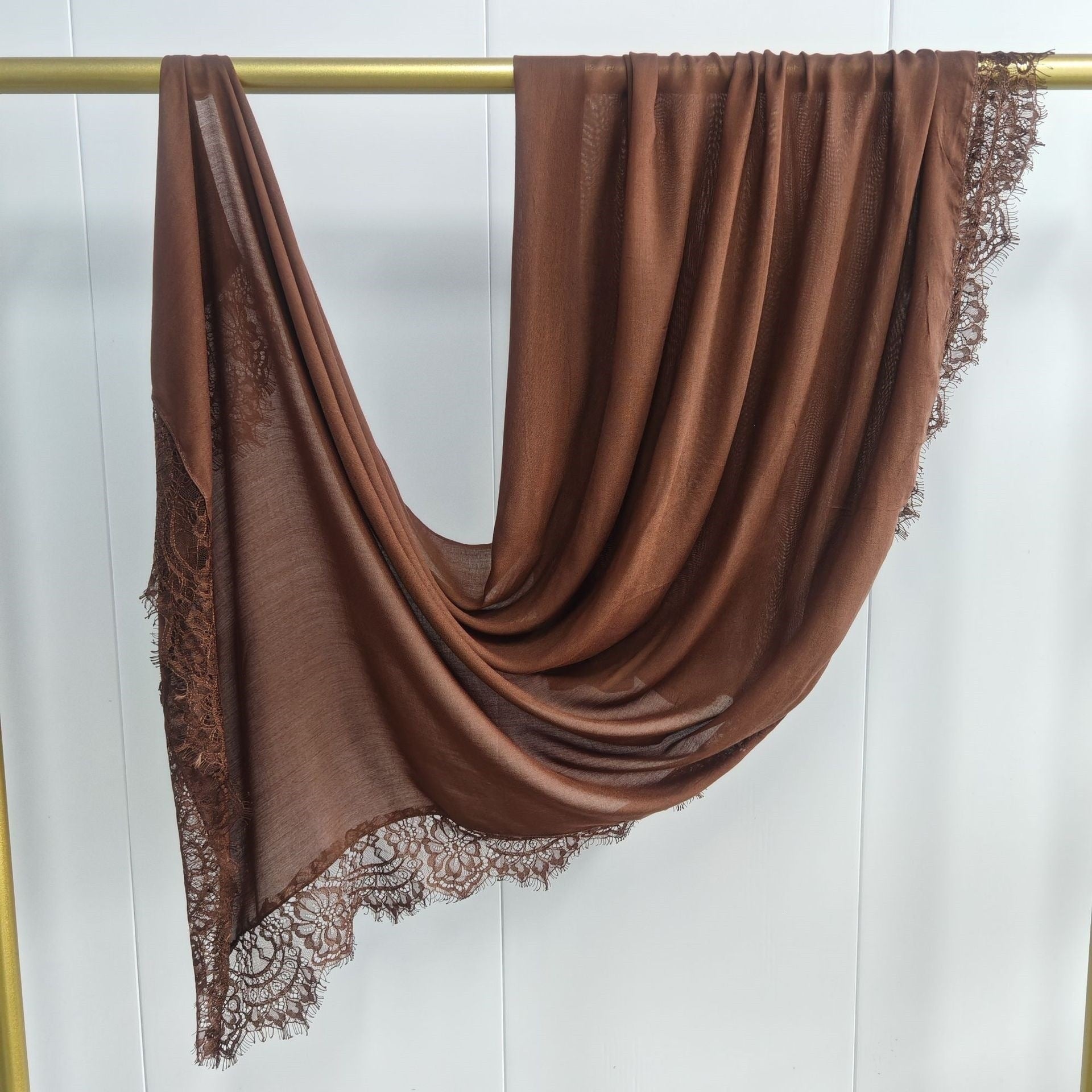 Brown scarf with lace trim draped over a gold rod on a white background