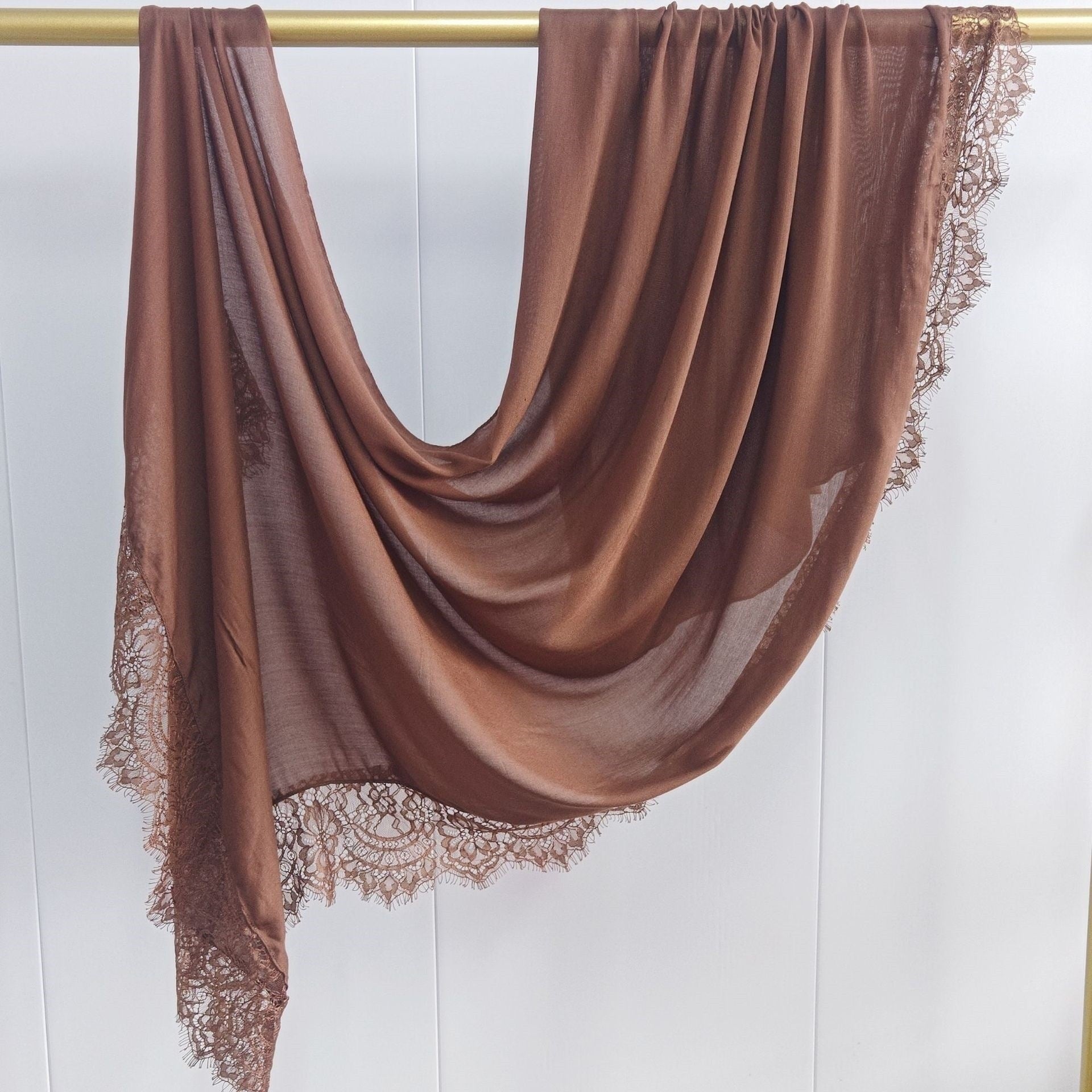 Brown scarf with lace trim draped over a gold rod on a white background