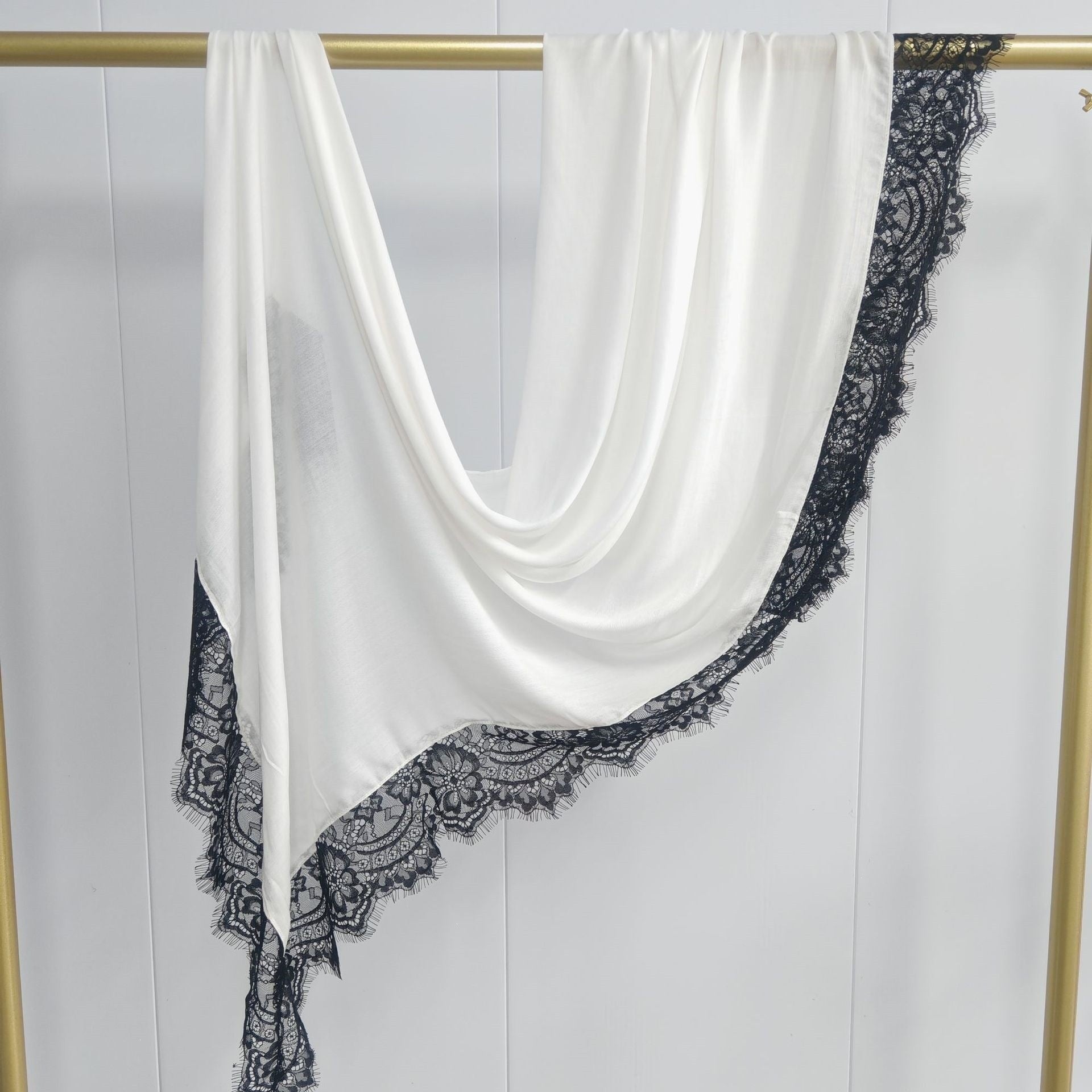 White scarf with black lace trim draped over a gold rod on a white background