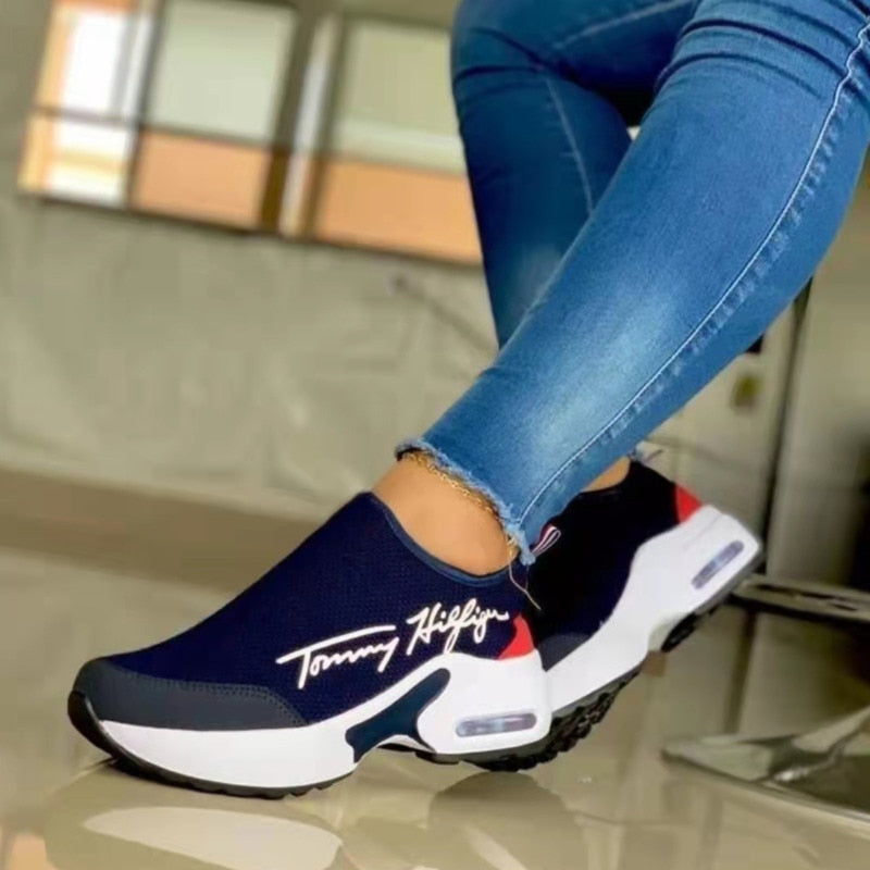 Navy and white slip-on shoes with red accents worn with blue jeans indoors.
