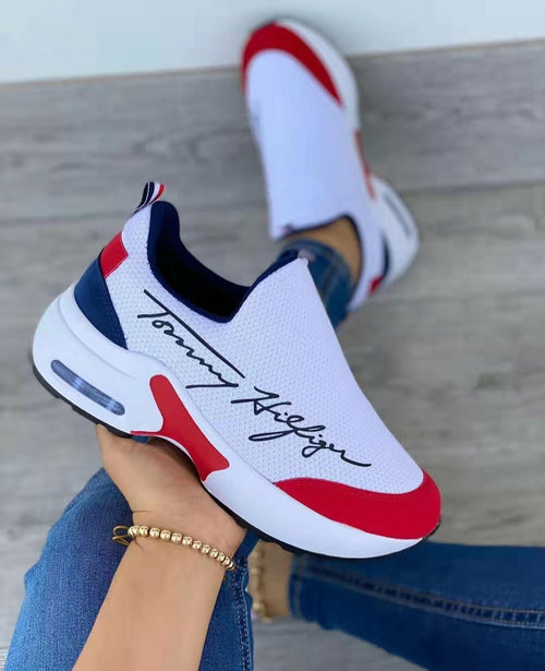 White sneaker with red and blue accents worn by a person sitting on a wooden floor.