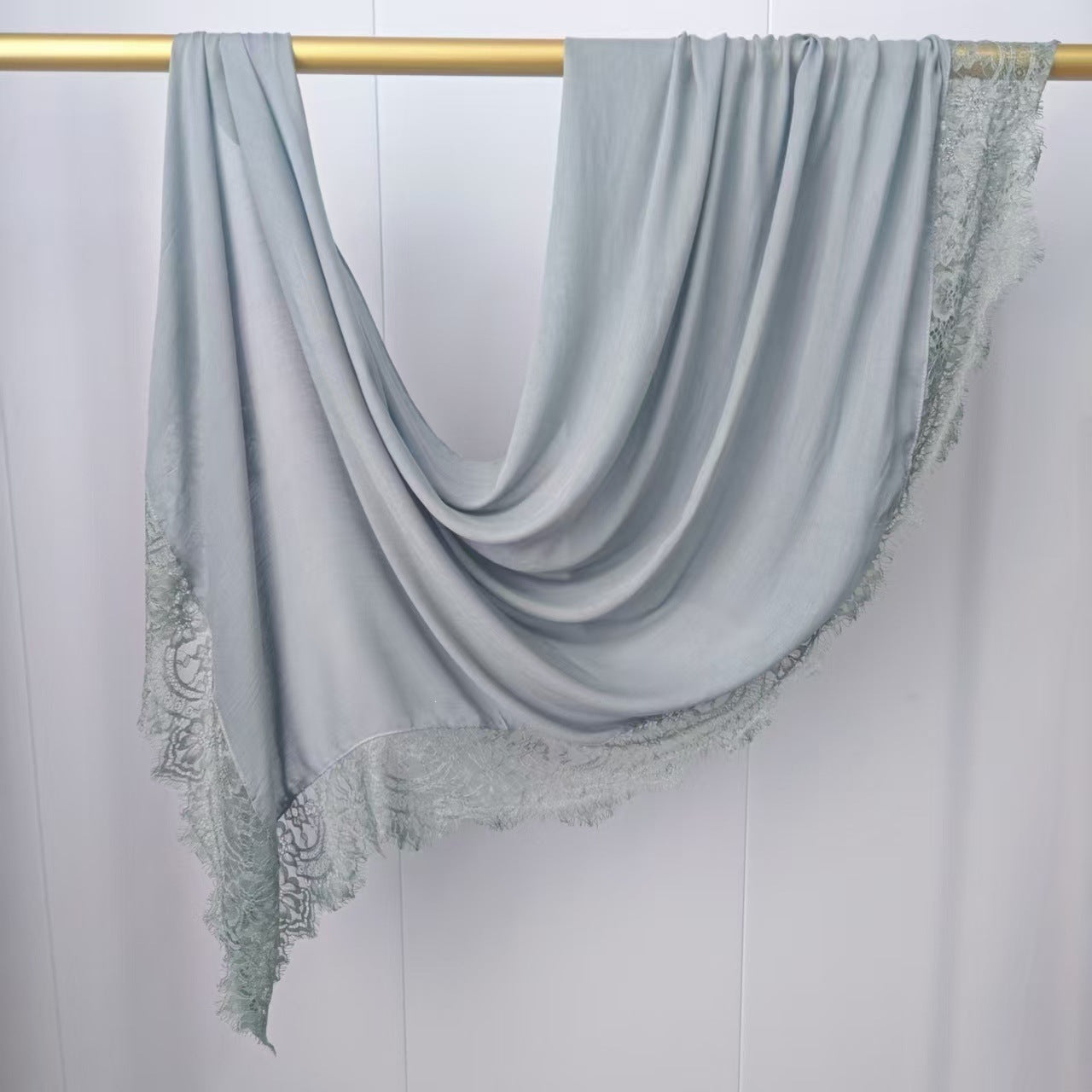 A light blue-gray square scarf with intricate lace trim along three edges, draped elegantly over a gold-colored horizontal rod against a white paneled wall.
