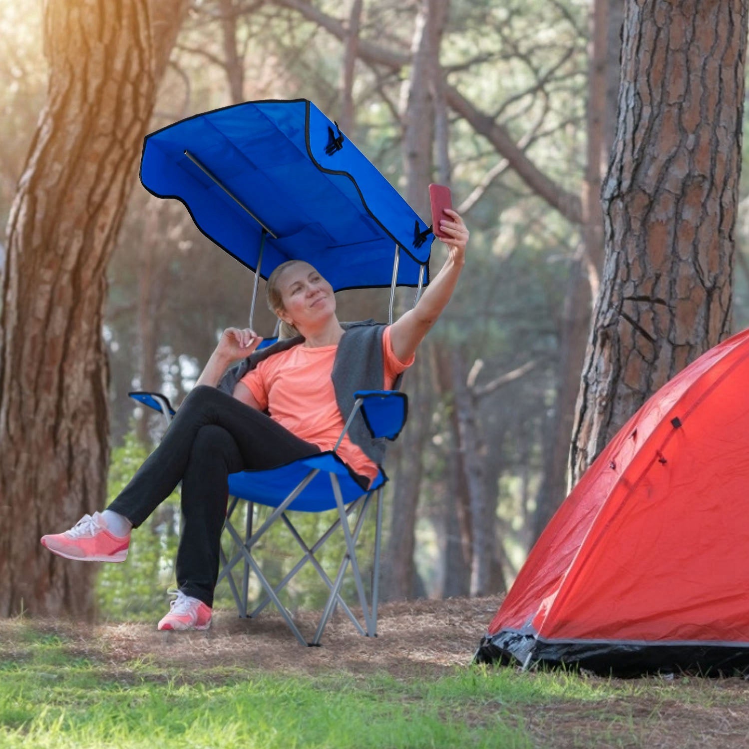 Person sitting in a camping chair with an attached canopy, taking a selfie in a forest setting with a red tent nearby.