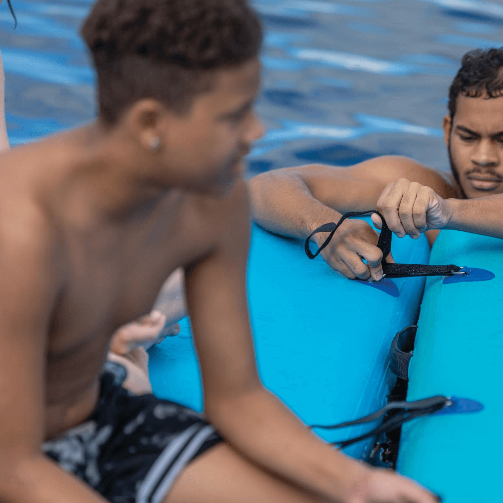 Two men sitting on a blue inflatable raft by a pool.