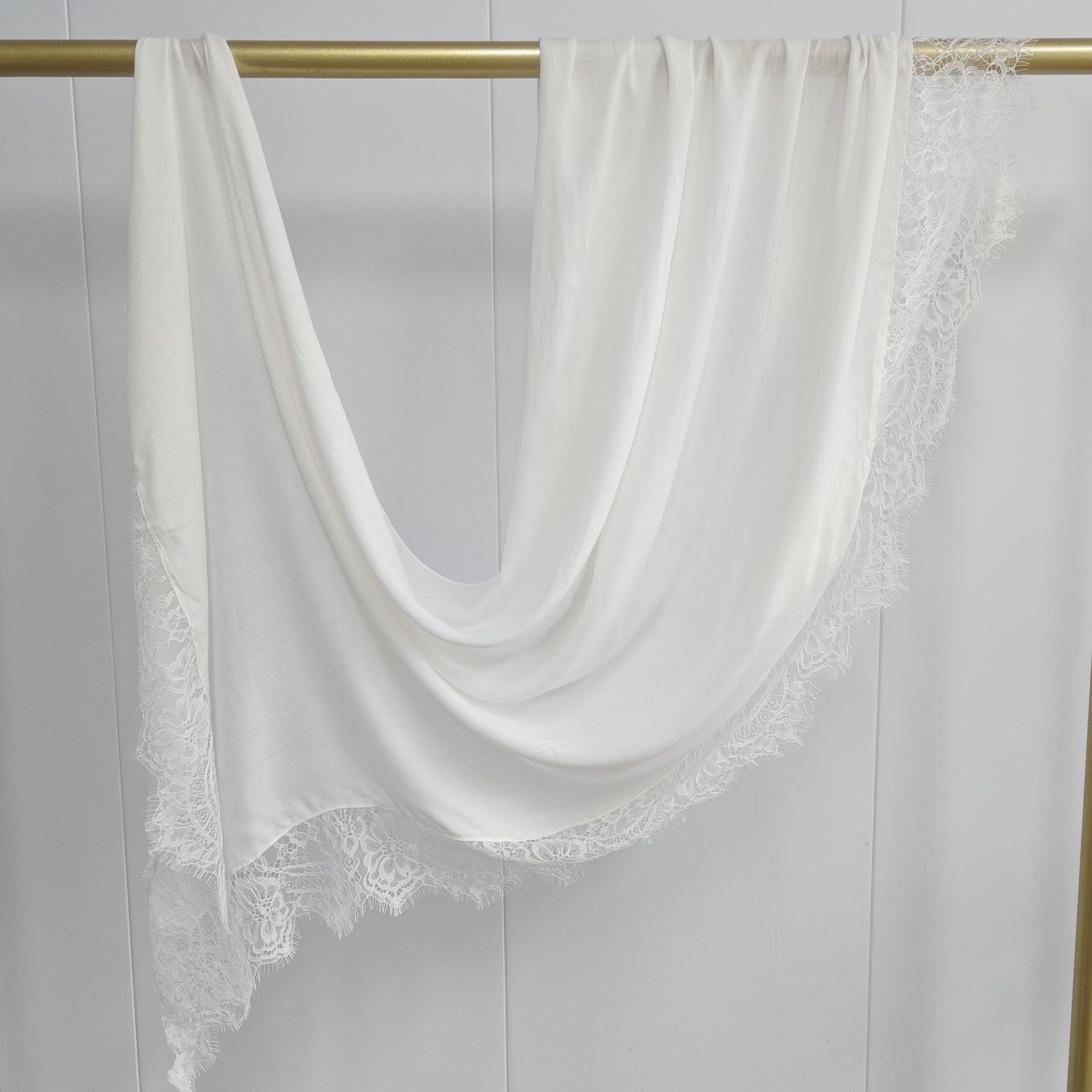 White scarf with lace trim draped over a gold rod against a gray background