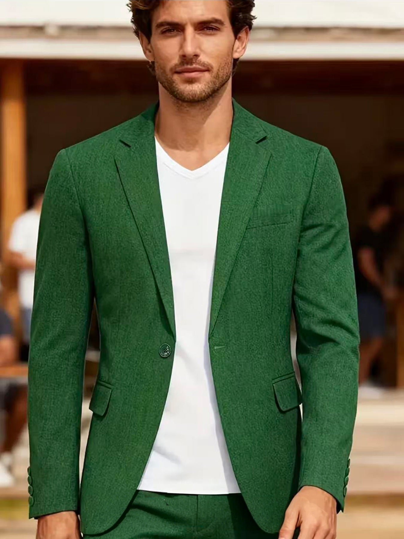 A man wearing a textured green blazer over a white V-neck t-shirt, standing in an outdoor setting with a blurred background.