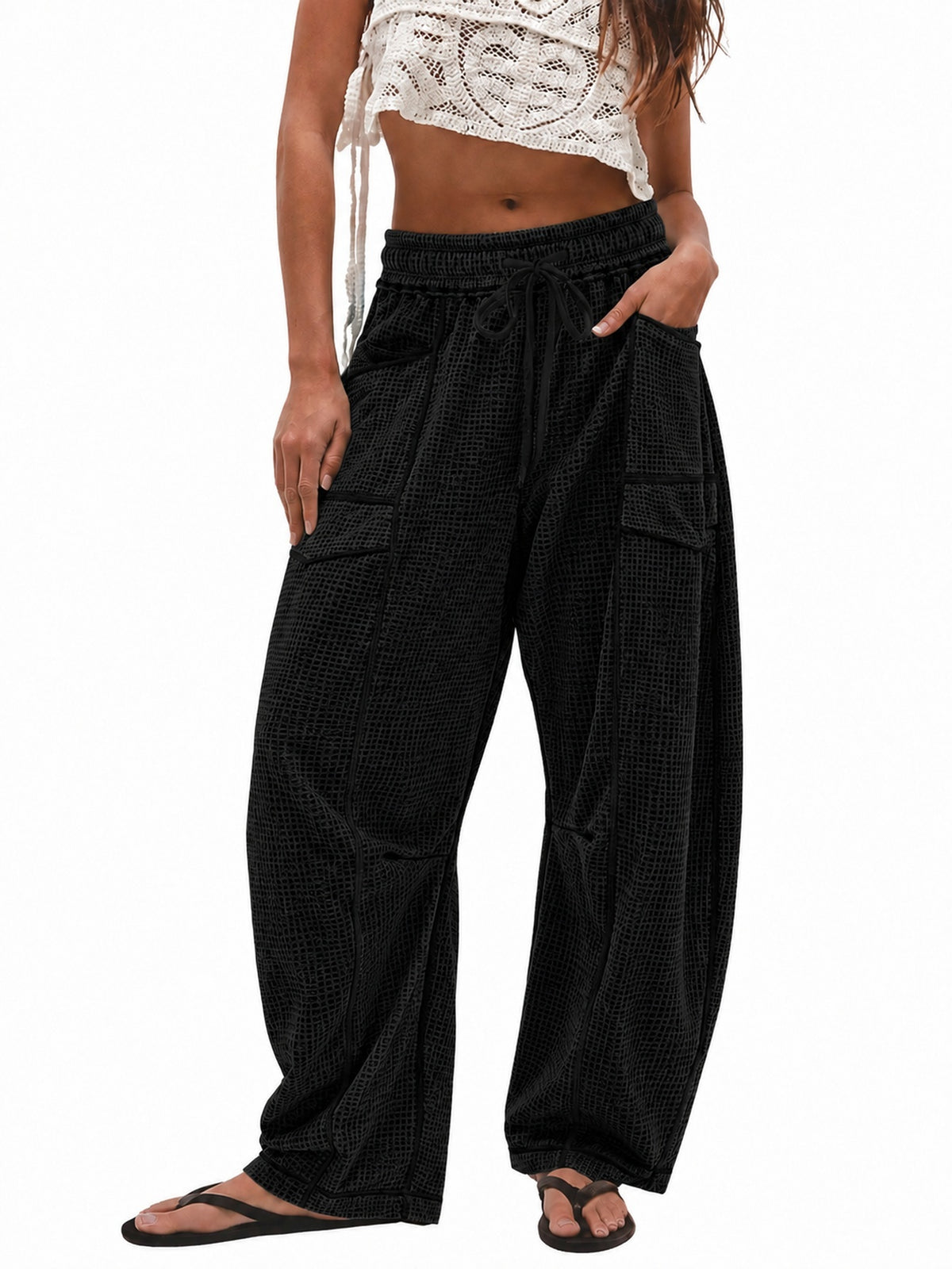 A model wearing black wide-leg cargo pants with a textured pattern, paired with a white crochet crop top and black flip-flop sandals, shown against a plain white background.