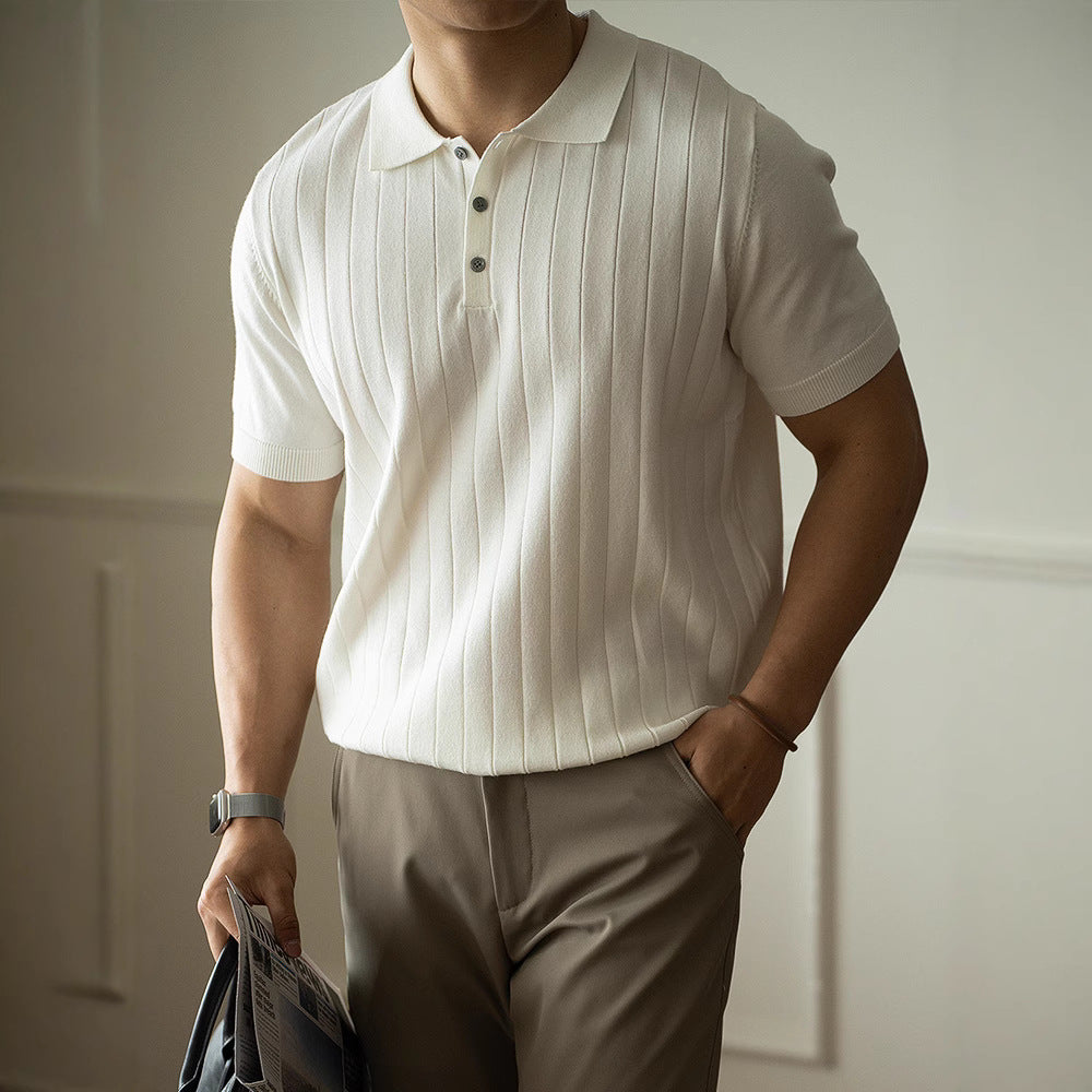 Man wearing a white ribbed polo shirt and beige pants, holding a newspaper indoors.