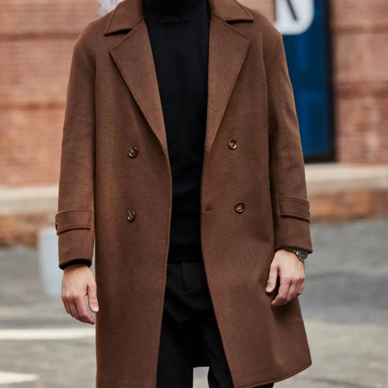 A man wearing a mid-length brown double-breasted overcoat over a black turtleneck sweater and black trousers, standing outdoors in front of a blurred brick building.