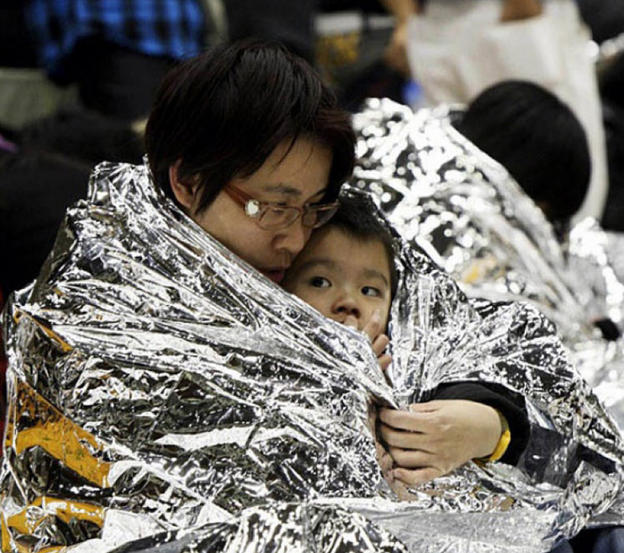 Two people wrapped in a reflective thermal blanket in an indoor setting.