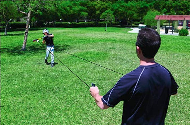 Two people playing baseball swing trainer a green course with trees in the background