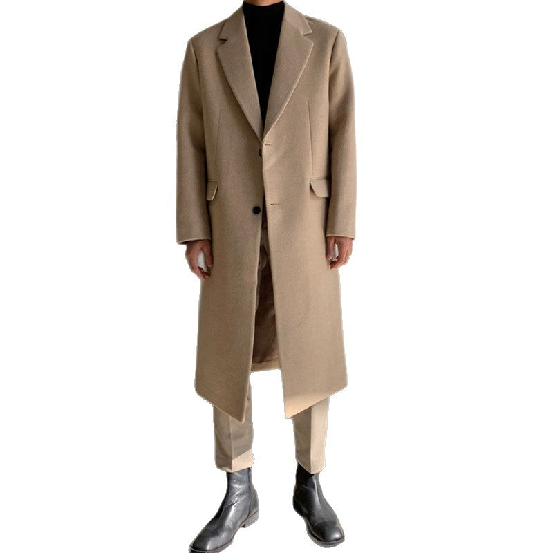 A model wearing a long, double-breasted camel-colored overcoat over a black turtleneck, matching beige trousers, and black leather boots, set against a plain white background.
