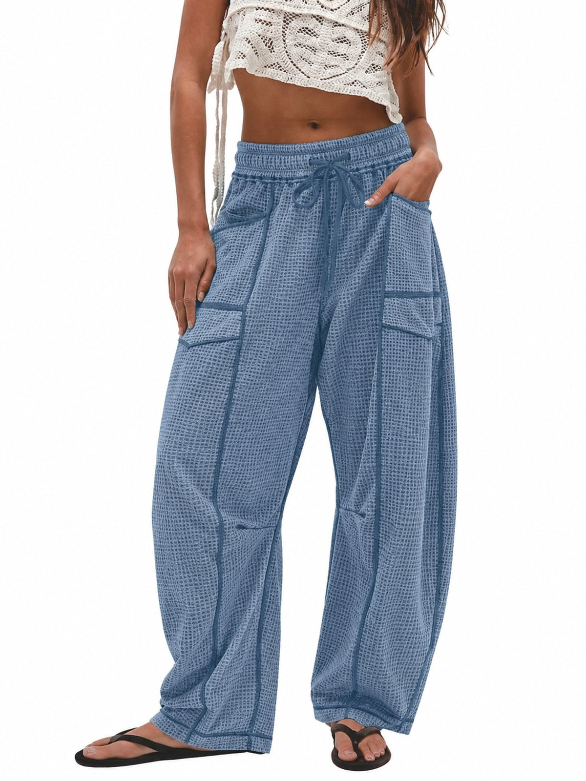 A model wearing blue patterned wide-leg harem pants with a drawstring waist and side cargo pockets, paired with a white crochet crop top and black flip-flops, shown against a plain white background.