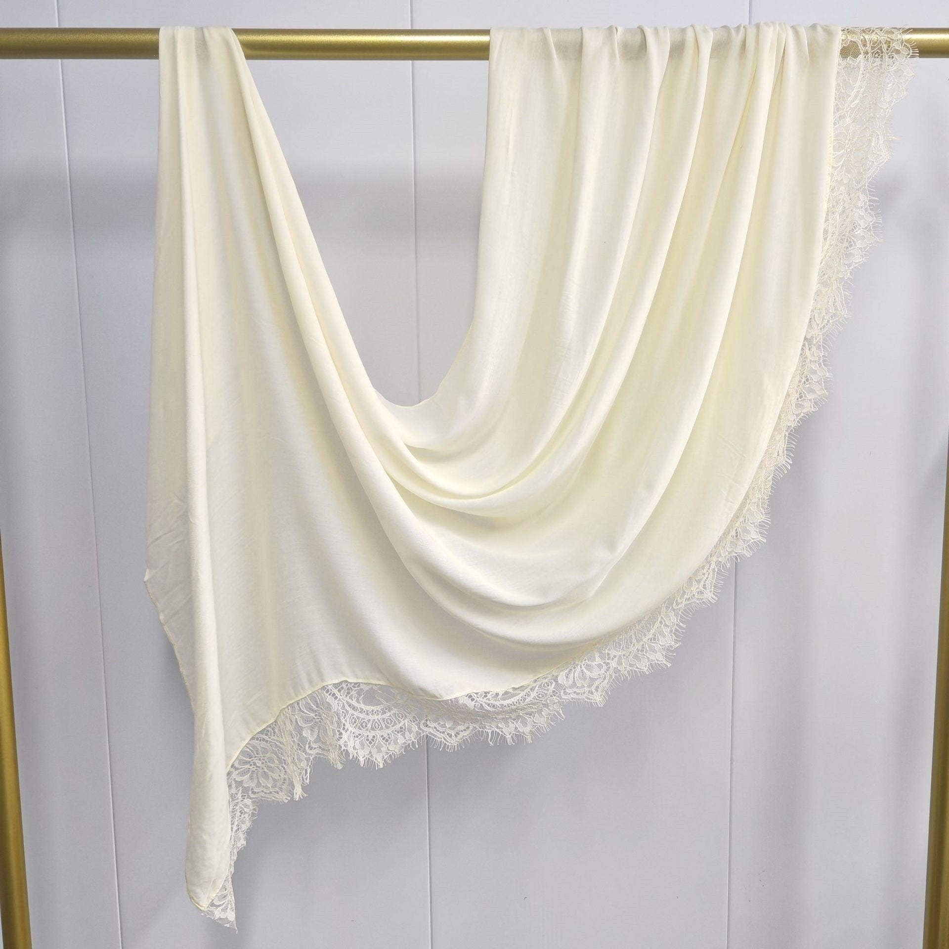 Cream-colored scarf with lace trim draped over a gold rod against a white background