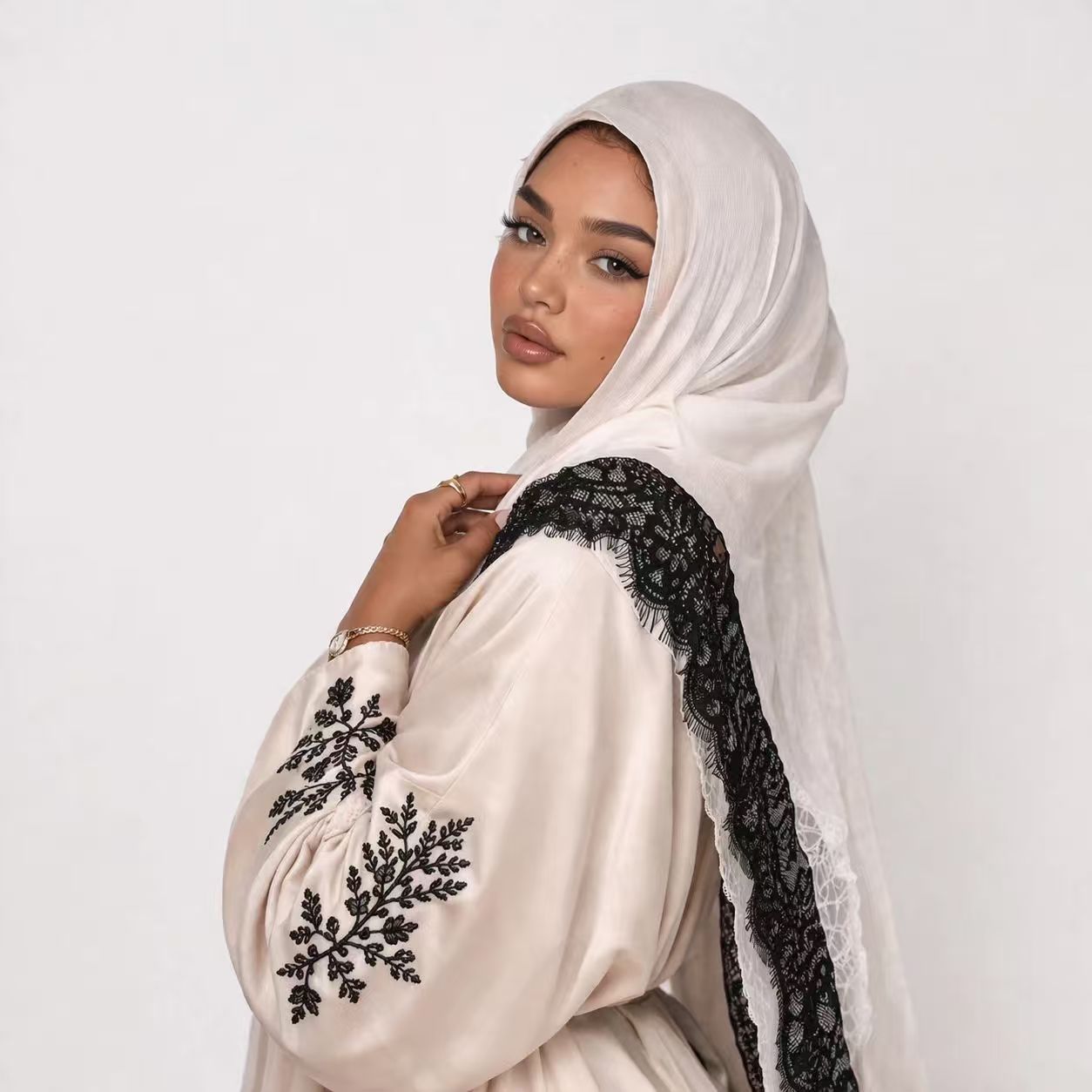 Woman wearing a white hijab and a white outfit with black embroidery on a plain background