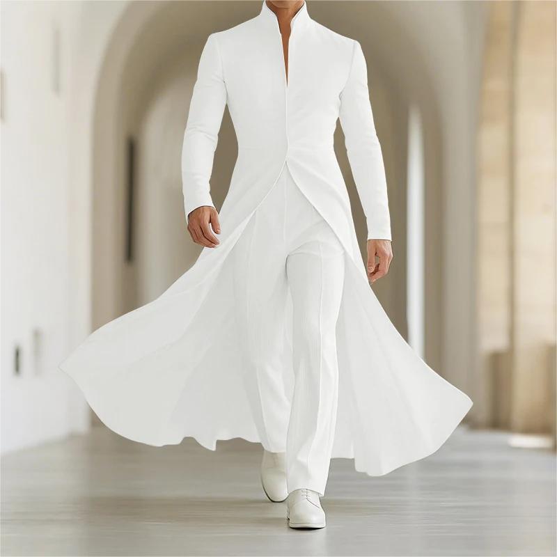 A model walks forward in a long white tailored outfit with a high collar, long sleeves, draped front panels, matching wide-leg trousers, and white shoes in a softly lit indoor corridor with arches.