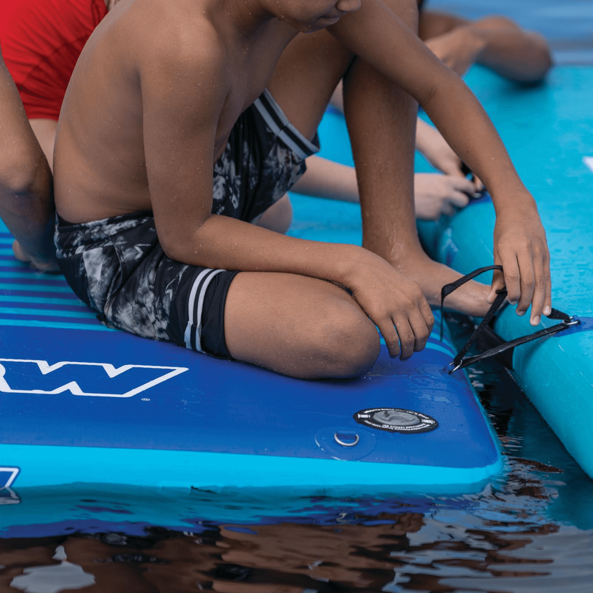 Children sitting on a blue Wavestorm surfboard in a pool.
