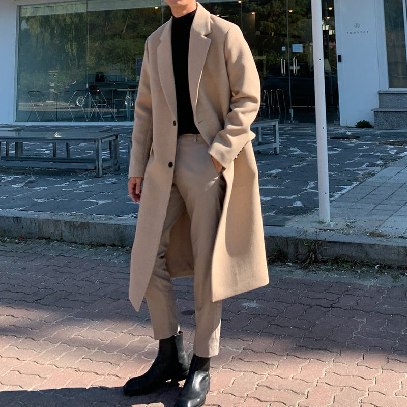 A man wearing a long beige overcoat, matching beige trousers, a black turtleneck, and black leather boots, standing outdoors on a paved street in front of a modern glass storefront.