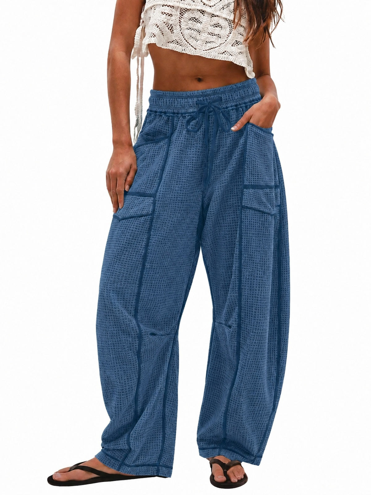 A model wearing blue wide-leg harem pants with a subtle grid pattern, side pockets, and a drawstring waist, paired with a white crochet crop top and black flip-flops against a white background.