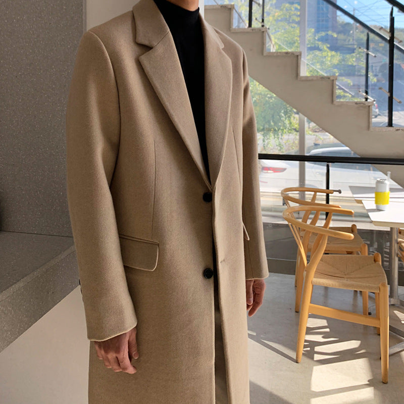 A person wearing a long beige overcoat over a black turtleneck, standing in a modern indoor setting with a staircase and wooden chairs visible in the background.