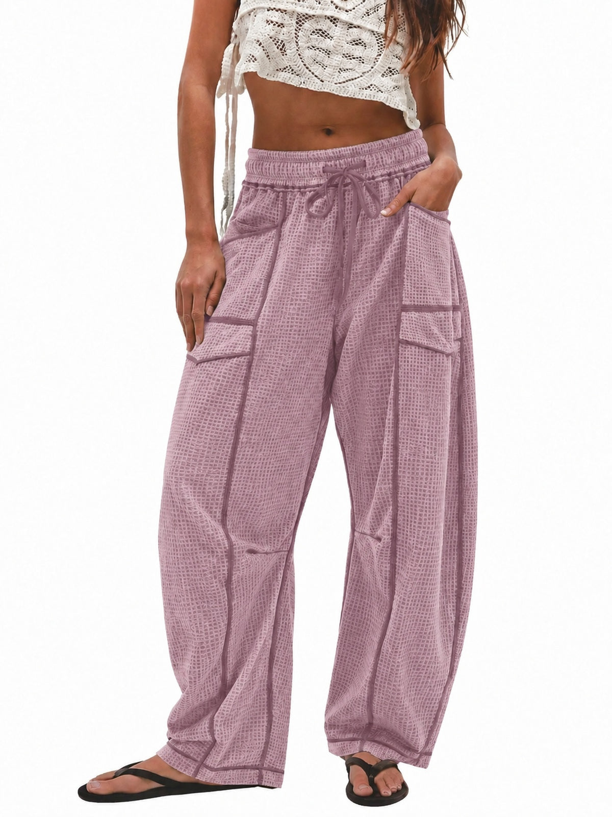 A model wearing loose, wide-leg lavender pants with a subtle grid pattern, side pockets, and an elastic drawstring waist, paired with a white crochet crop top and black flip-flops against a plain white background.