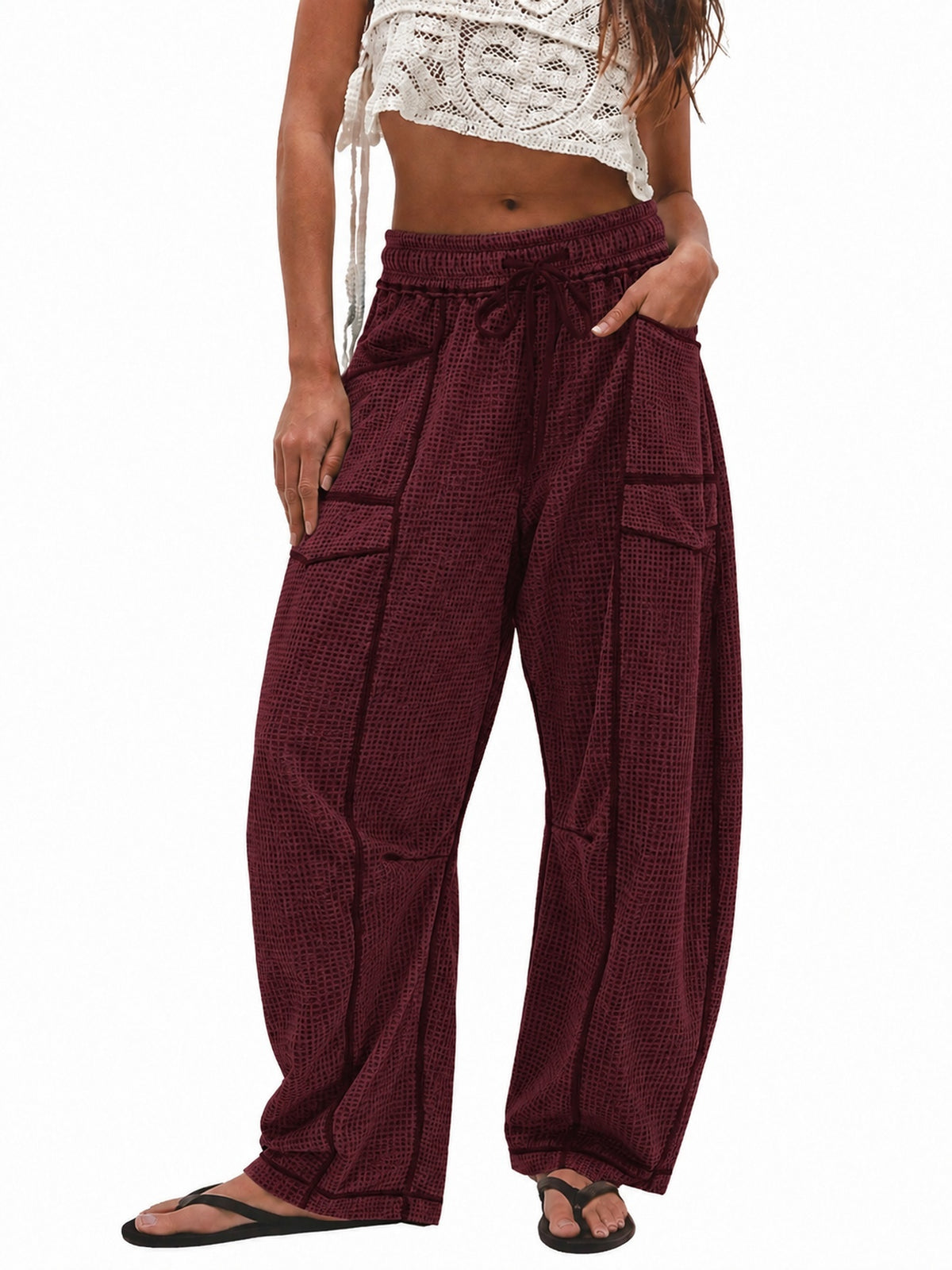 A model wearing loose, wide-leg harem-style pants in a deep burgundy color with a subtle grid pattern. The pants feature an elastic drawstring waist, side pockets, and large cargo-style pockets on the thighs, paired with black flip-flop sandals against a plain white background.