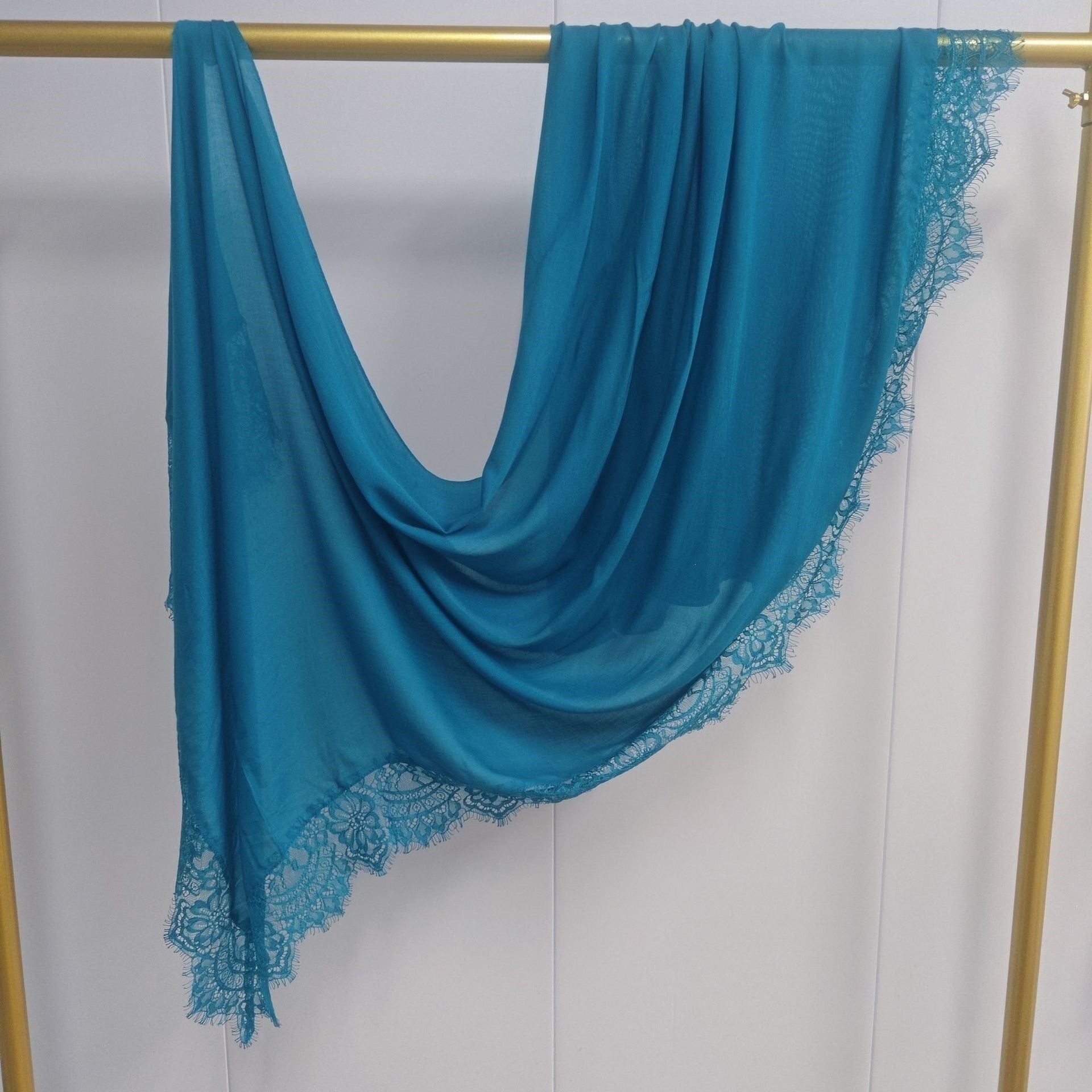 Turquoise scarf with lace trim draped over a gold rod against a white background