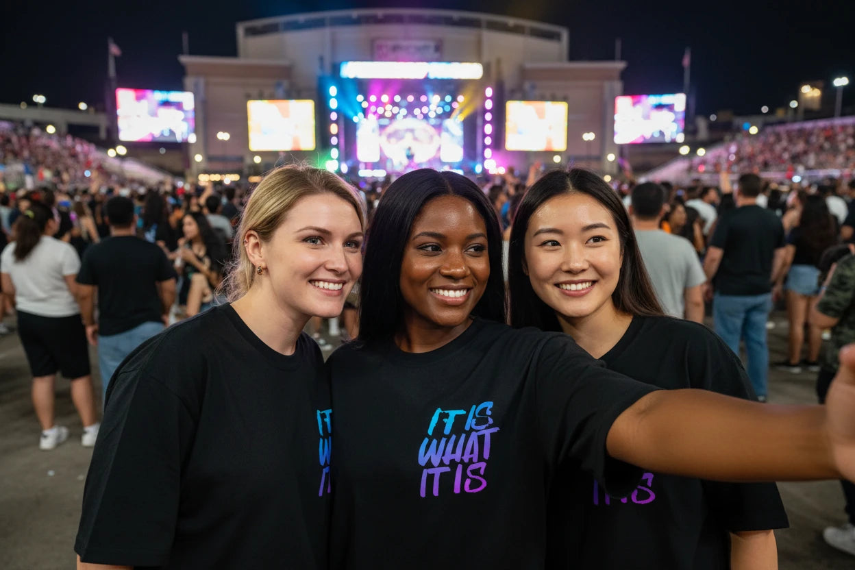 Black t-shirt with colorful text 'IT IS WHAT IT IS' worn by a three people outdoor concert