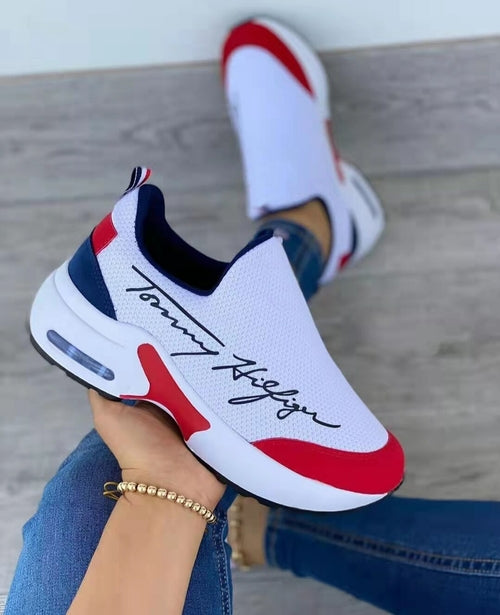 White sneaker with red and blue accents worn by a person sitting on a wooden floor.