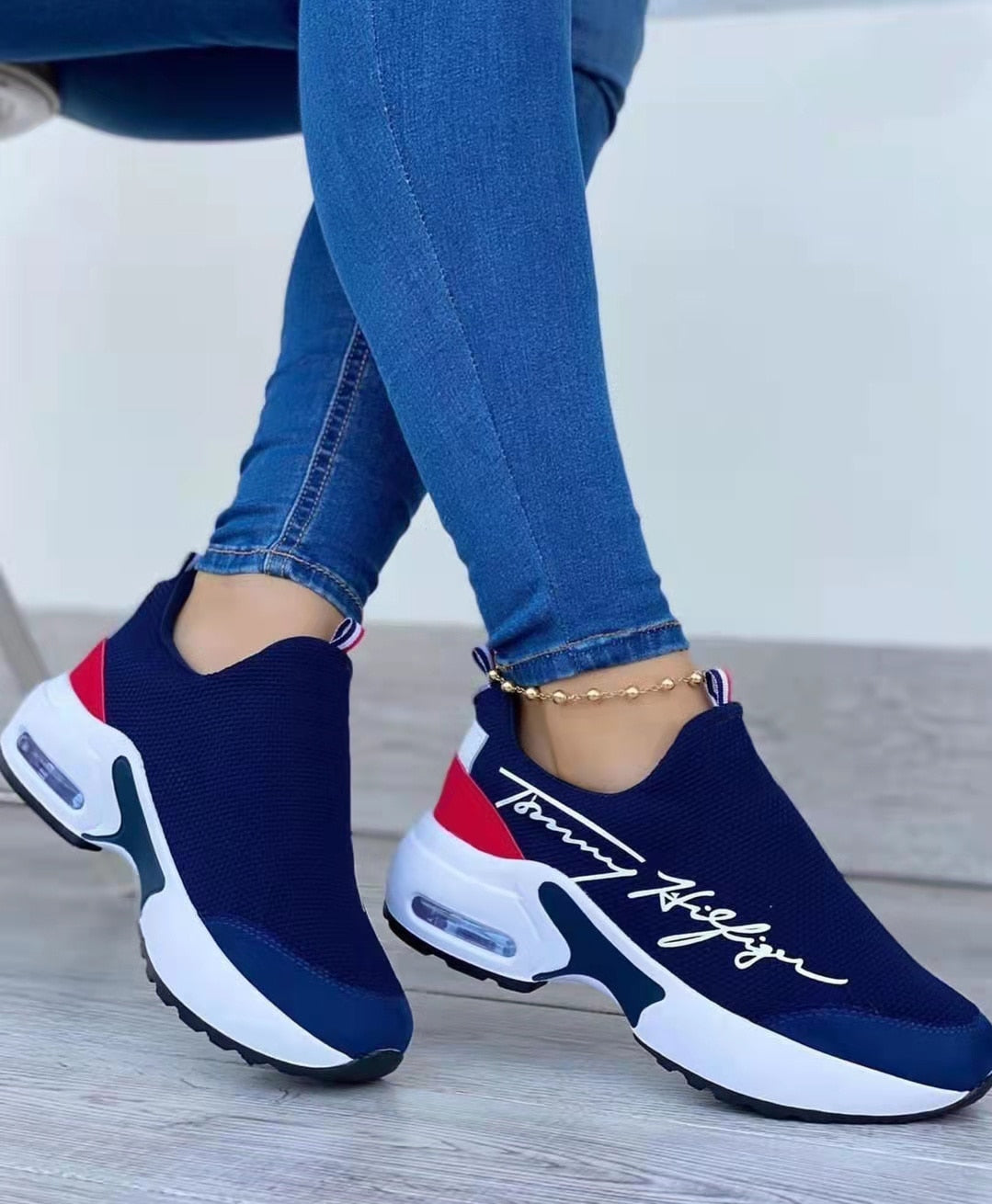 Navy slip-on sneakers with red and white accents worn with blue jeans on a light wooden floor.