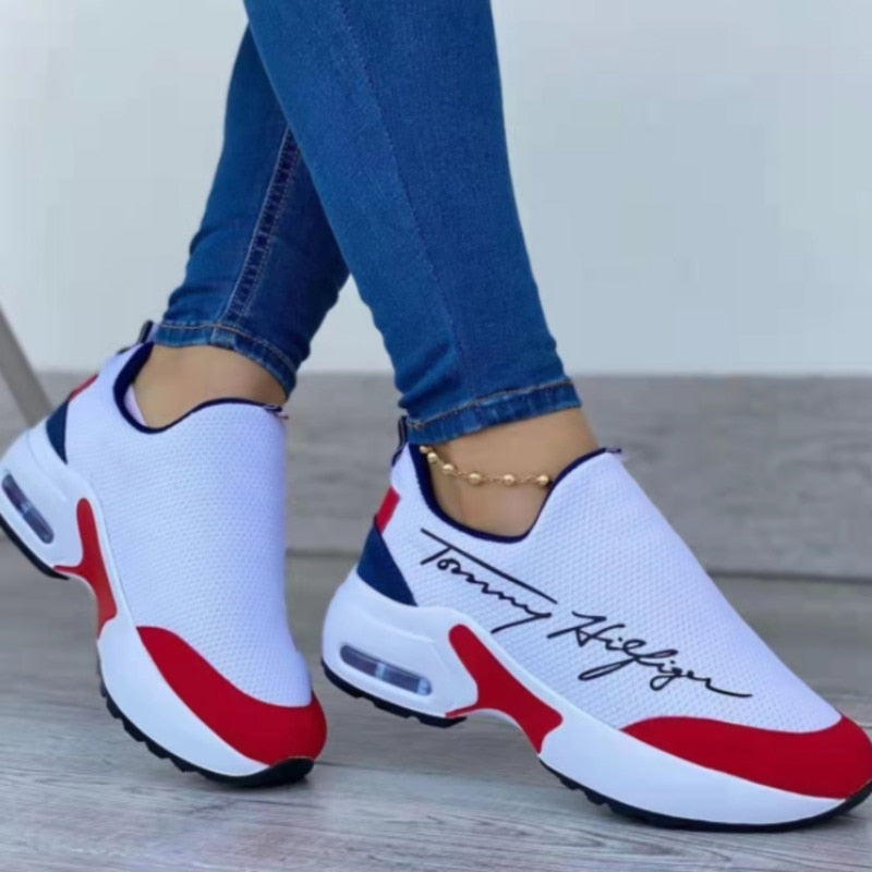 White sneakers with red and blue accents worn with blue jeans on a light wooden floor.