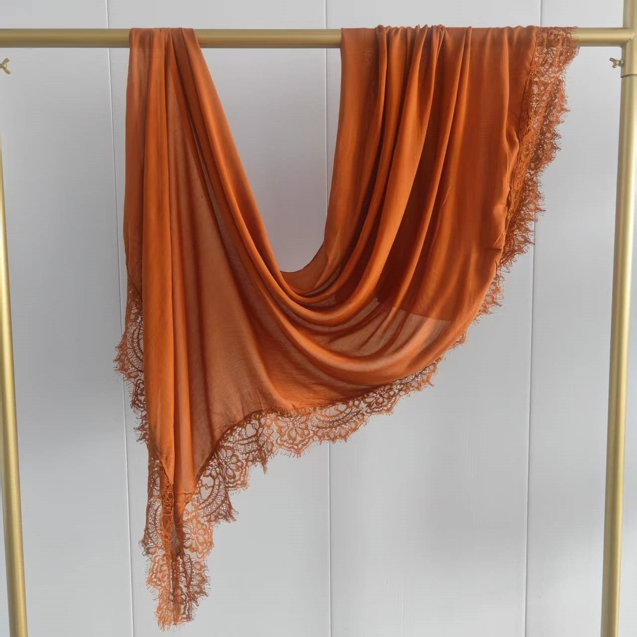 An orange draped scarf with a delicate lace border hanging from a gold clothing rack against a white paneled wall.