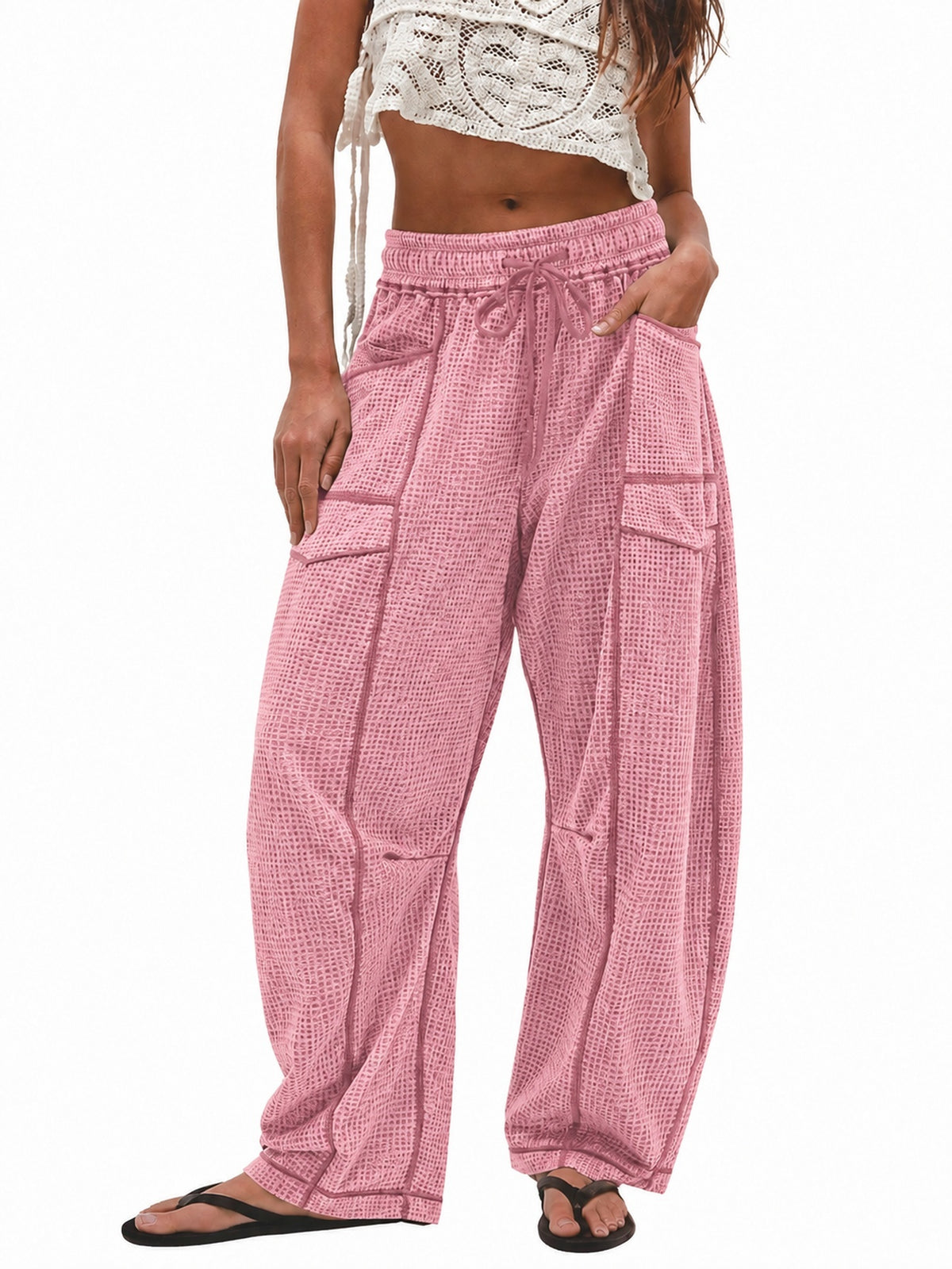 A model wearing pink and white checkered harem-style pants with a drawstring waist, side cargo pockets, and a relaxed baggy fit, paired with a white crochet crop top and black flip-flop sandals against a plain white background.