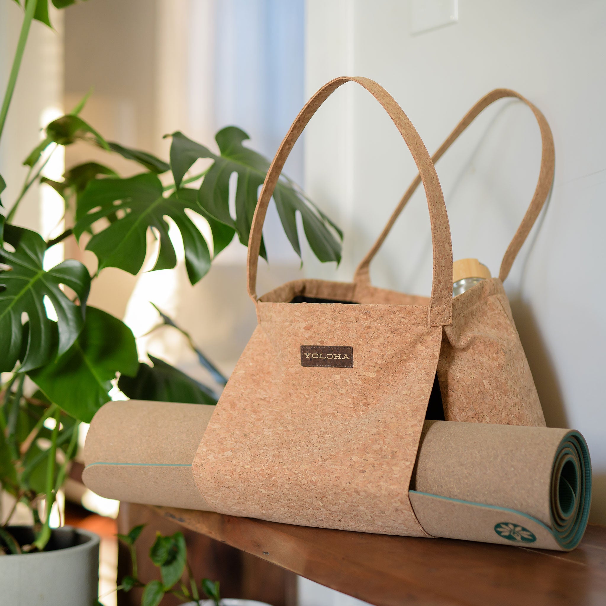 Cork tote bag with rolled cork mat on a wooden surface, surrounded by plants.